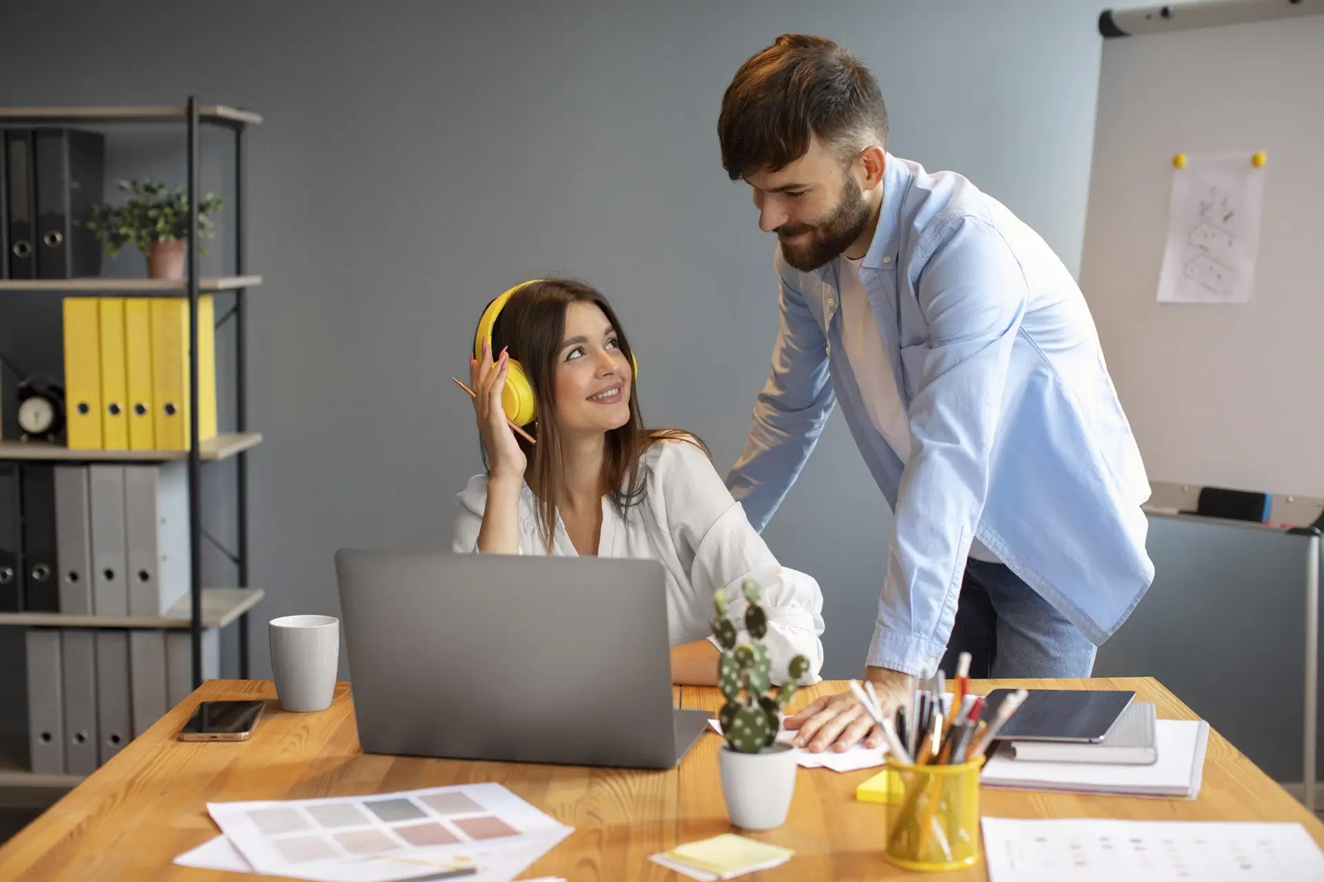 Mulher com fone e notebook conversando com homem em pé de camisa clara em ambiente de escritório
